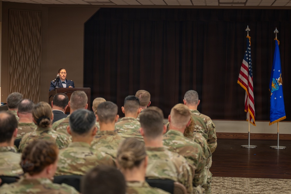 A military officer in dress uniform is delivering a speech at a podium to an audience of uniformed personnel seated in rows. The setting appears to be a formal military ceremony or briefing held in an indoor venue. Two flags — the United States flag and what appears to be the U.S. Air Force flag — are displayed on stands to the right of the stage.