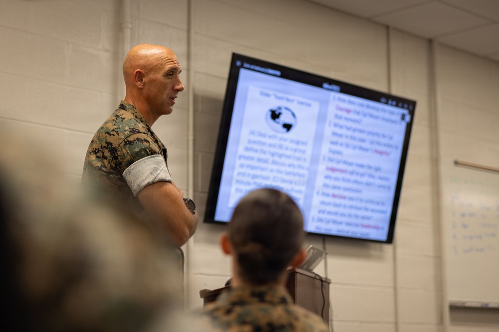 A bald male military personnel in Marine Corps MARPAT camouflage uniform stands with arms crossed in what appears to be a classroom or briefing room. He is presenting or instructing in front of a large display screen showing text content, while at least one other uniformed individual is seated facing the screen. The setting suggests a formal military training or educational session.