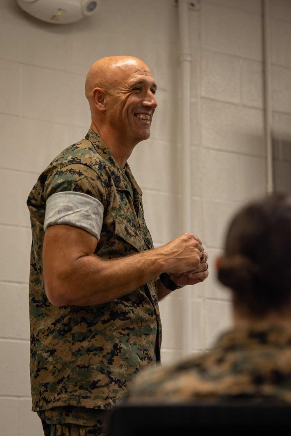 A bald, muscular male military personnel wearing MARPAT (Marine pattern) camouflage uniform is standing and smiling broadly, appearing to address an audience. He has his hands clasped together in front of him and is wearing a black wristband/watch. At least one other uniformed individual is visible in the foreground, out of focus.