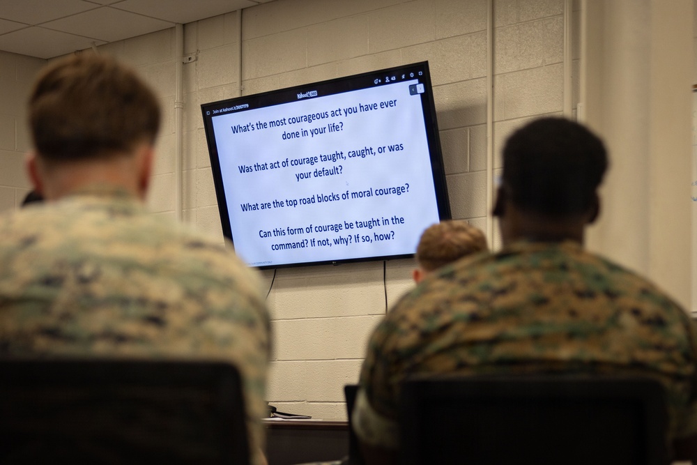 Military personnel in camouflage uniforms are seated in a classroom setting, viewing a presentation on a flat-screen monitor. The screen displays discussion questions related to moral courage, suggesting a leadership or ethics training session. The platform visible on the screen appears to be Kahoot, an interactive learning tool.