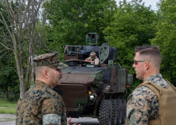 The image shows U.S. Marines in woodland MARPAT camouflage uniforms observing a wheeled armored fighting vehicle, likely an ACV (Amphibious Combat Vehicle) or similar 8x8 wheeled platform. A crew member is visible in the open hatch of the vehicle. The setting appears to be a paved area on a military installation surrounded by trees.