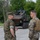 The image shows U.S. Marines in woodland MARPAT camouflage uniforms observing a wheeled armored fighting vehicle, likely an ACV (Amphibious Combat Vehicle) or similar 8x8 wheeled platform. A crew member is visible in the open hatch of the vehicle. The setting appears to be a paved area on a military installation surrounded by trees.