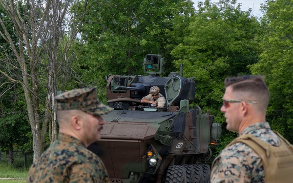 The image shows U.S. Marines in woodland MARPAT camouflage uniforms observing a wheeled armored fighting vehicle, likely an ACV (Amphibious Combat Vehicle) or similar 8x8 wheeled platform. A crew member is visible in the open hatch of the vehicle. The setting appears to be a paved area on a military installation surrounded by trees.