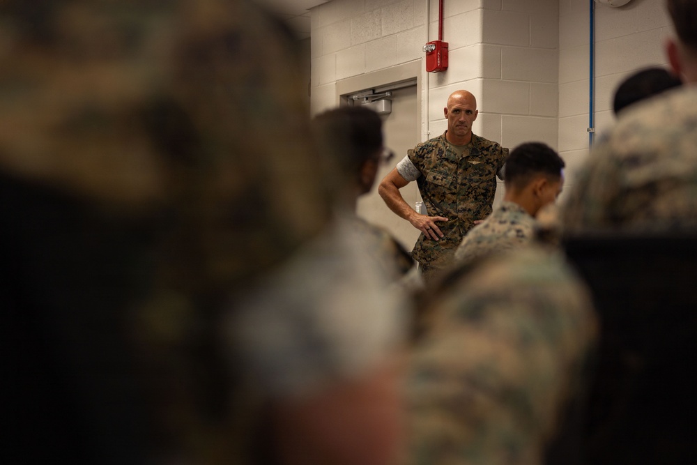 A bald military officer in woodland camouflage uniform stands with hands on hips, appearing to address a group of seated or gathered service members in an indoor facility. The scene suggests a briefing, instruction, or command address taking place in what appears to be a military building. The foreground figures are intentionally blurred, drawing focus to the standing officer in the background.