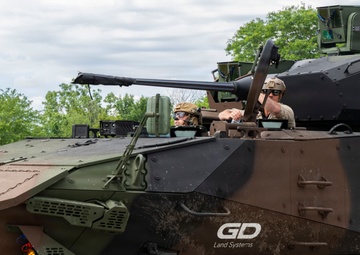 The image shows a General Dynamics Land Systems (GDLS) wheeled armored fighting vehicle, likely a Stryker variant or similar 8x8 armored personnel carrier, in a multi-tone camouflage paint scheme. Two personnel wearing military helmets and eye protection are visible in open hatches. The vehicle is photographed outdoors in a green, vegetated environment, likely during a demonstration or exercise.