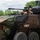 The image shows a General Dynamics Land Systems (GDLS) wheeled armored fighting vehicle, likely a Stryker variant or similar 8x8 armored personnel carrier, in a multi-tone camouflage paint scheme. Two personnel wearing military helmets and eye protection are visible in open hatches. The vehicle is photographed outdoors in a green, vegetated environment, likely during a demonstration or exercise.