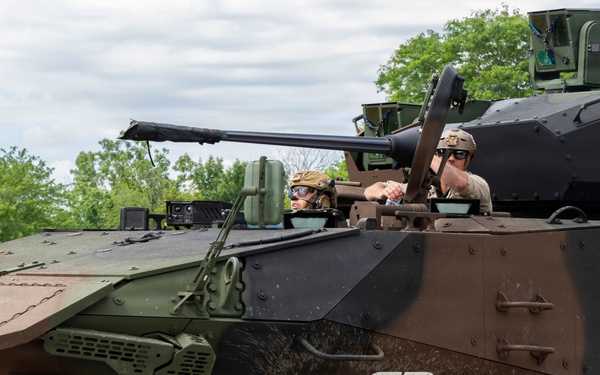 The image shows a General Dynamics Land Systems (GDLS) wheeled armored fighting vehicle, likely a Stryker variant or similar 8x8 armored personnel carrier, in a multi-tone camouflage paint scheme. Two personnel wearing military helmets and eye protection are visible in open hatches. The vehicle is photographed outdoors in a green, vegetated environment, likely during a demonstration or exercise.