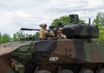 An 8-wheeled armored fighting vehicle manufactured by General Dynamics Land Systems (GDLS) is shown on a paved surface in a wooded area. The vehicle displays a three-tone camouflage pattern of green, brown, and black, with two personnel visible in the open top hatches. The vehicle appears to be at a demonstration or evaluation event based on its clean presentation and the 'GD Land Systems' branding visible on the hull.