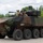 An 8-wheeled armored fighting vehicle manufactured by General Dynamics Land Systems (GDLS) is shown on a paved surface in a wooded area. The vehicle displays a three-tone camouflage pattern of green, brown, and black, with two personnel visible in the open top hatches. The vehicle appears to be at a demonstration or evaluation event based on its clean presentation and the 'GD Land Systems' branding visible on the hull.