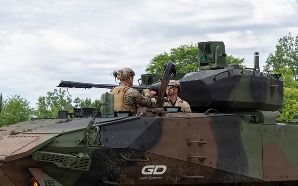 An 8-wheeled armored fighting vehicle manufactured by General Dynamics Land Systems (GDLS) is shown on a paved surface in a wooded area. The vehicle displays a three-tone camouflage pattern of green, brown, and black, with two personnel visible in the open top hatches. The vehicle appears to be at a demonstration or evaluation event based on its clean presentation and the 'GD Land Systems' branding visible on the hull.