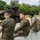 A group of U.S. Marines in digital camouflage uniforms are seated along a concrete barrier observing a light armored vehicle (LAV) or similar wheeled armored fighting vehicle during what appears to be a training or demonstration exercise. The vehicle, painted in a brown and green camouflage pattern, has a crew member visible in the turret. The setting appears to be a military installation or training area with trees visible in the background.