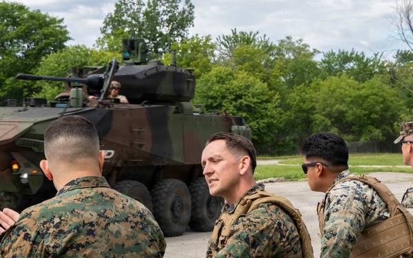 A group of U.S. Marines in digital camouflage uniforms are seated along a concrete barrier observing a light armored vehicle (LAV) or similar wheeled armored fighting vehicle during what appears to be a training or demonstration exercise. The vehicle, painted in a brown and green camouflage pattern, has a crew member visible in the turret. The setting appears to be a military installation or training area with trees visible in the background.