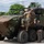 An 8-wheeled armored combat vehicle, consistent with a Marine Personnel Carrier (MPC) or similar amphibious combat vehicle, is shown in an outdoor paved area surrounded by trees. Two individuals in military camouflage uniforms are positioned on or near the top hatch of the vehicle, while a civilian in a black shirt and khaki pants stands beside it holding what appears to be a green-handled tool or implement. The scene appears to be a demonstration, inspection, or familiarization event.