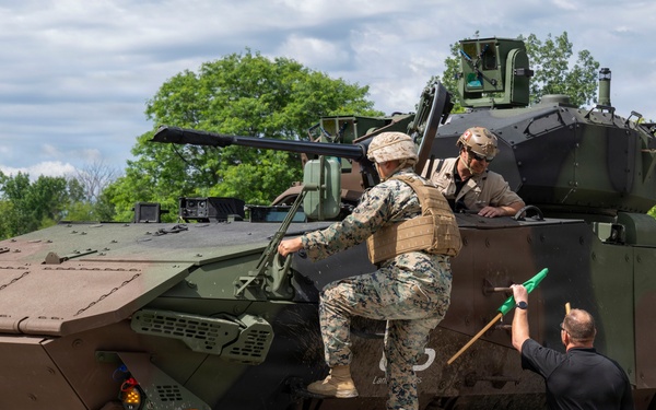 An 8-wheeled armored combat vehicle, consistent with a Marine Personnel Carrier (MPC) or similar amphibious combat vehicle, is shown in an outdoor paved area surrounded by trees. Two individuals in military camouflage uniforms are positioned on or near the top hatch of the vehicle, while a civilian in a black shirt and khaki pants stands beside it holding what appears to be a green-handled tool or implement. The scene appears to be a demonstration, inspection, or familiarization event.