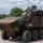 An 8-wheeled armored combat vehicle manufactured by General Dynamics Land Systems (GDLS) is shown operating on a paved surface in a green, tree-lined environment. Two personnel are visible in the open hatch area of the vehicle, which features a camouflage paint scheme in brown and green tones. The vehicle appears to be undergoing a demonstration or evaluation exercise.