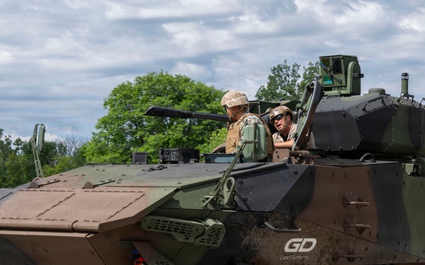 An 8-wheeled armored combat vehicle manufactured by General Dynamics Land Systems (GDLS) is shown operating on a paved surface in a green, tree-lined environment. Two personnel are visible in the open hatch area of the vehicle, which features a camouflage paint scheme in brown and green tones. The vehicle appears to be undergoing a demonstration or evaluation exercise.