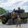 Two military personnel in camouflage uniforms and tactical gear are interacting with a large 8-wheeled armored combat vehicle in an outdoor setting. One soldier is climbing onto the vehicle while another stands nearby observing. The vehicle appears to be a modern amphibious combat vehicle with a turret-mounted weapon system, painted in a green and brown camouflage pattern.