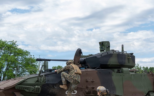 Two military personnel in camouflage uniforms and tactical gear are interacting with a large 8-wheeled armored combat vehicle in an outdoor setting. One soldier is climbing onto the vehicle while another stands nearby observing. The vehicle appears to be a modern amphibious combat vehicle with a turret-mounted weapon system, painted in a green and brown camouflage pattern.