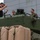A civilian in casual clothing appears to be briefing or demonstrating something to three military personnel in camouflage uniforms atop what appears to be a green armored military vehicle. Several tan/beige jerry cans labeled 'FUEL' and 'WATER' are visible in the foreground. The setting appears to be an outdoor area near a modern building.