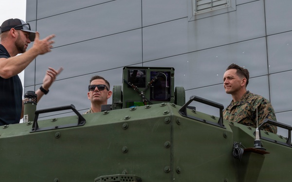 A civilian in casual clothing appears to be briefing or demonstrating something to three military personnel in camouflage uniforms atop what appears to be a green armored military vehicle. Several tan/beige jerry cans labeled 'FUEL' and 'WATER' are visible in the foreground. The setting appears to be an outdoor area near a modern building.
