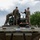 Several military personnel in camouflage uniforms are gathered on top of and around a modern armored military vehicle, appearing to conduct an inspection or familiarization exercise. One individual in civilian attire (dark shirt and cap) appears to be briefing or demonstrating something to the uniformed personnel. The setting appears to be a military installation or base, with trees and overcast skies in the background.