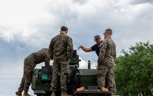 Several military personnel in camouflage uniforms are gathered on top of and around a modern armored military vehicle, appearing to conduct an inspection or familiarization exercise. One individual in civilian attire (dark shirt and cap) appears to be briefing or demonstrating something to the uniformed personnel. The setting appears to be a military installation or base, with trees and overcast skies in the background.