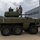 Three individuals, two in military uniforms and one in civilian clothing, are standing atop a large wheeled armored military vehicle with a turret and cannon. The vehicle appears to be an 8-wheeled armored fighting vehicle painted in olive drab green, positioned on a concrete surface next to a modern building. A metal access staircase is positioned against the right side of the vehicle.