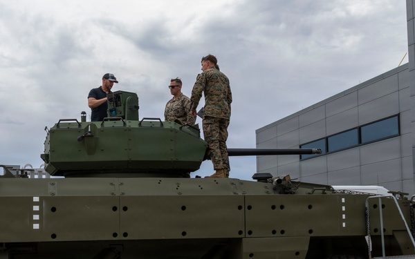 Three individuals, two in military uniforms and one in civilian clothing, are standing atop a large wheeled armored military vehicle with a turret and cannon. The vehicle appears to be an 8-wheeled armored fighting vehicle painted in olive drab green, positioned on a concrete surface next to a modern building. A metal access staircase is positioned against the right side of the vehicle.