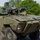 A modern 8-wheeled armored fighting vehicle (AFV) painted in olive/military green is photographed in motion on a paved surface. The vehicle is equipped with a turret-mounted cannon and has a crew member visible in an open hatch. The setting appears to be a military testing or demonstration area surrounded by trees.