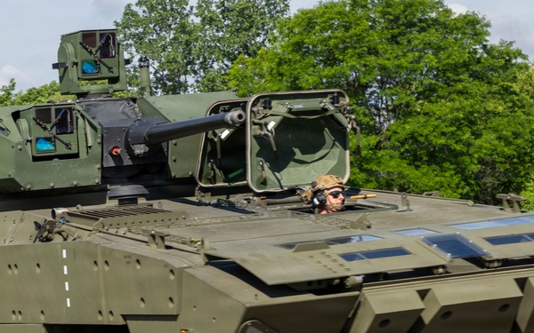 A modern 8-wheeled armored fighting vehicle (AFV) painted in olive/military green is photographed in motion on a paved surface. The vehicle is equipped with a turret-mounted cannon and has a crew member visible in an open hatch. The setting appears to be a military testing or demonstration area surrounded by trees.