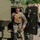 A U.S. Marine in full combat uniform and tactical vest is exiting or demonstrating a military vehicle, likely during a public display or demonstration event. A civilian man in a black jacket observes and interacts near the vehicle door. The setting appears to be an outdoor military equipment exhibition with trees visible in the background.