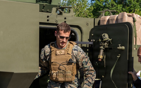A U.S. Marine in full combat uniform and tactical vest is exiting or demonstrating a military vehicle, likely during a public display or demonstration event. A civilian man in a black jacket observes and interacts near the vehicle door. The setting appears to be an outdoor military equipment exhibition with trees visible in the background.
