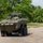 A military wheeled armored personnel carrier or infantry fighting vehicle in olive green livery is being driven on a paved road course marked with orange traffic cones. A crew member is visible in the open hatch wearing a helmet and protective gear. The setting appears to be a training or demonstration exercise conducted in a grassy, tree-lined area.
