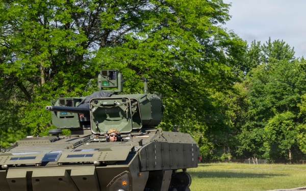 A military wheeled armored personnel carrier or infantry fighting vehicle in olive green livery is being driven on a paved road course marked with orange traffic cones. A crew member is visible in the open hatch wearing a helmet and protective gear. The setting appears to be a training or demonstration exercise conducted in a grassy, tree-lined area.