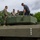 Several military personnel in camouflage uniforms and at least one civilian are gathered on top of a large military armored vehicle, examining its turret. The vehicle appears to be a modern armored fighting vehicle painted in olive drab green. The setting appears to be an outdoor demonstration or familiarization event.