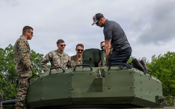 Several military personnel in camouflage uniforms and at least one civilian are gathered on top of a large military armored vehicle, examining its turret. The vehicle appears to be a modern armored fighting vehicle painted in olive drab green. The setting appears to be an outdoor demonstration or familiarization event.