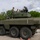 A large 8-wheeled armored military vehicle, painted in olive drab green, is parked on a paved surface surrounded by personnel in military camouflage uniforms and at least one civilian. Several individuals are positioned on top of the vehicle near the turret, appearing to conduct an inspection or demonstration. The setting appears to be an outdoor military evaluation or demonstration event.