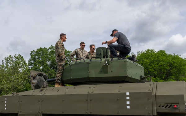 A large 8-wheeled armored military vehicle, painted in olive drab green, is parked on a paved surface surrounded by personnel in military camouflage uniforms and at least one civilian. Several individuals are positioned on top of the vehicle near the turret, appearing to conduct an inspection or demonstration. The setting appears to be an outdoor military evaluation or demonstration event.