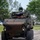 A large wheeled armored military vehicle is being driven on a paved road in what appears to be a military installation or demonstration area. Two personnel are visible in the open hatch/turret area of the vehicle. The setting includes green trees, grass, and civilian vehicles visible in the background.