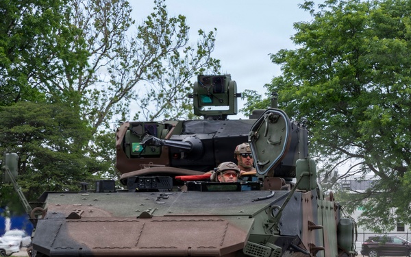 A large wheeled armored military vehicle is being driven on a paved road in what appears to be a military installation or demonstration area. Two personnel are visible in the open hatch/turret area of the vehicle. The setting includes green trees, grass, and civilian vehicles visible in the background.