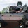 A male operator wearing a combat helmet and sunglasses is seated in the open hatch of a modern armored military vehicle, appearing to be driving or operating it. The vehicle is a wheeled armored fighting vehicle with a remote weapon station mounted on top, featuring a two-tone brown and olive green camouflage scheme. A 'GD' (General Dynamics) logo is partially visible on the lower right side of the hull.