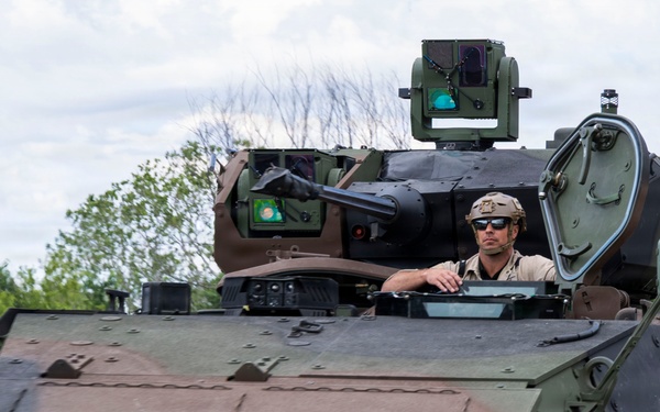 A male operator wearing a combat helmet and sunglasses is seated in the open hatch of a modern armored military vehicle, appearing to be driving or operating it. The vehicle is a wheeled armored fighting vehicle with a remote weapon station mounted on top, featuring a two-tone brown and olive green camouflage scheme. A 'GD' (General Dynamics) logo is partially visible on the lower right side of the hull.