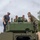 Four individuals — one civilian and three military personnel in woodland camouflage uniforms — are gathered on top of a green military vehicle, examining or working on a mounted equipment unit. The setting appears to be an outdoor field environment with trees and an overcast sky. The scene suggests a technical demonstration, inspection, or equipment installation activity.