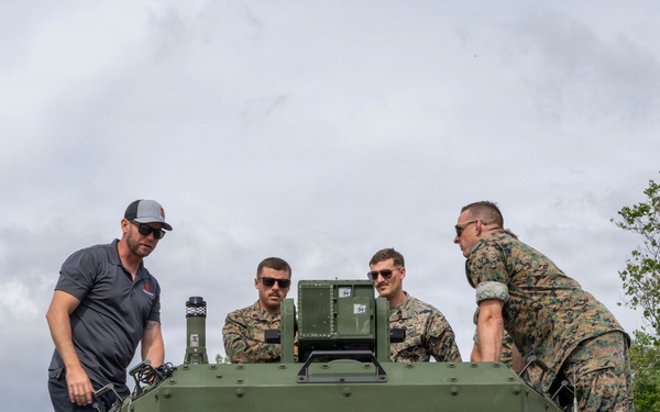 Four individuals — one civilian and three military personnel in woodland camouflage uniforms — are gathered on top of a green military vehicle, examining or working on a mounted equipment unit. The setting appears to be an outdoor field environment with trees and an overcast sky. The scene suggests a technical demonstration, inspection, or equipment installation activity.