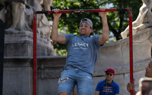 A young man wearing a gray 'Legacy Road Trip' t-shirt and ripped denim shorts is performing pull-ups on a red metal bar frame in an outdoor public space. A woman in the foreground is recording him on a smartphone, while another person sits on steps in the background. The setting appears to be near a monument or classical stone sculpture, suggesting an urban park or plaza.