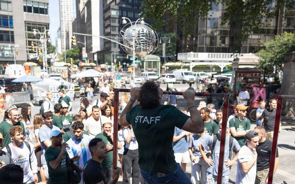 An outdoor public fitness event is taking place in what appears to be Columbus Circle in New York City, identifiable by the globe sculpture in the background. A person wearing a 'STAFF' t-shirt is performing pull-ups on a red portable pull-up bar frame while a crowd of onlookers watches. Several participants wear matching green shirts, and a person in a U.S. Marines t-shirt reading 'Pain is Weakness Leaving the Body' is visible in the foreground.