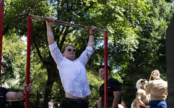 A civilian man in a white button-up shirt and black pants is performing pull-ups on a red pull-up bar at what appears to be a U.S. Marines public outreach or recruiting event. Two Marines in camouflage uniforms are standing nearby, one stabilizing the bar structure. In the background, additional Marines in tan dress uniforms appear to be part of a band setup.