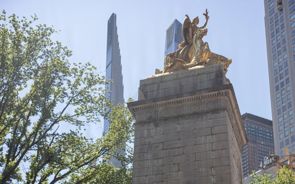 A U.S. Marine Corps band performs outdoors in front of a large stone monument topped with a gilded sculptural group, surrounded by trees and modern skyscrapers in the background. The musicians are dressed in standard Marine Corps service uniforms (tan blouses, blue trousers with red stripe) and are playing a variety of brass, woodwind, and percussion instruments. The setting appears to be Columbus Circle or a similar urban plaza in New York City.
