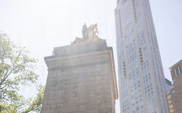 A U.S. Marines military band performs outdoors in an urban plaza, positioned in front of a large stone monument with sculptural figures at its apex. Musicians in tan/khaki service uniforms play various instruments including a sousaphone, saxophone, trombone, and drum kit. A tall modern skyscraper is visible in the background, suggesting a major city setting, likely New York City.