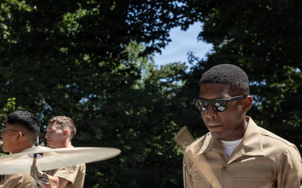 A group of U.S. Marine Corps musicians are performing outdoors, likely at a public event or parade. The focal point is a Marine playing a drum kit, wearing sunglasses and the standard tan and blue Marine Corps service uniform. Additional Marines are visible in the background, also in uniform, with one appearing to play cymbals.